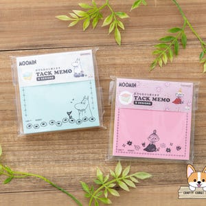 Könnte beinhalten: Zwei Haftnotizblöcke im Moomin-Design in Kunststoffverpackung. Einer ist hellblau, der andere rosa. Jeder Block hat eine andere Moomin-Charakterillustration und den Text "TACK MEMO 5 DESIGNS". Blumen- und Sternmuster umranden die Notizen.