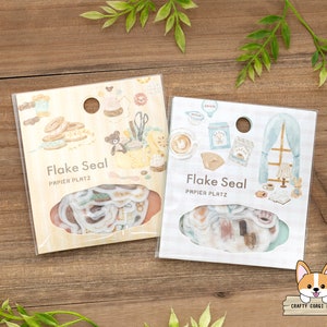 May include: Two packs of decorative paper flakes, each with a different theme. One pack features a light blue background with a window and a book, and the other pack features a light brown background with a sewing theme. Both packs have the text "Flake Seal Papier Platz" on the front.