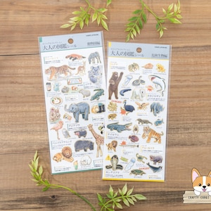 Puede incluir: Dos hojas de pegatinas con ilustraciones de animales y el texto "Guía ilustrada para adultos". Las pegatinas están sobre una superficie de madera.