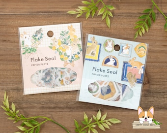 May include: Two packs of "Flake Seal" stickers featuring bunny illustrations. One pack has bunnies with floral arrangements, the other has bunnies in framed portraits. The stickers are visible through clear packaging.