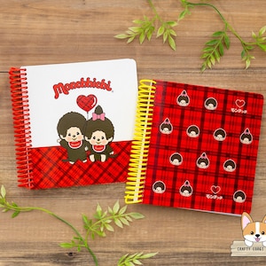 May include: Two spiral-bound notebooks with red and yellow coils rest on a wooden surface. One notebook features the text "Monchhichi" and a cartoon couple holding a heart balloon. The other has a red plaid pattern with cartoon faces.