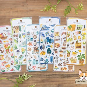 Puede incluir: Una colección de hojas de pegatinas decorativas con varios temas. Los diseños incluyen flores, casas, animales y artículos relacionados con el café. Cada hoja está empaquetada en una funda de plástico transparente con una cabecera amarilla. La marca "Quilt Atelier" está impresa.