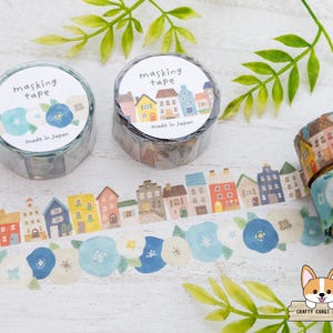 May include: Masking tape rolls with floral and house designs. One roll features blue flowers, another has colorful houses. The tape is unrolled, displaying the patterns. Green leaves and the text "masking tape" are also visible.