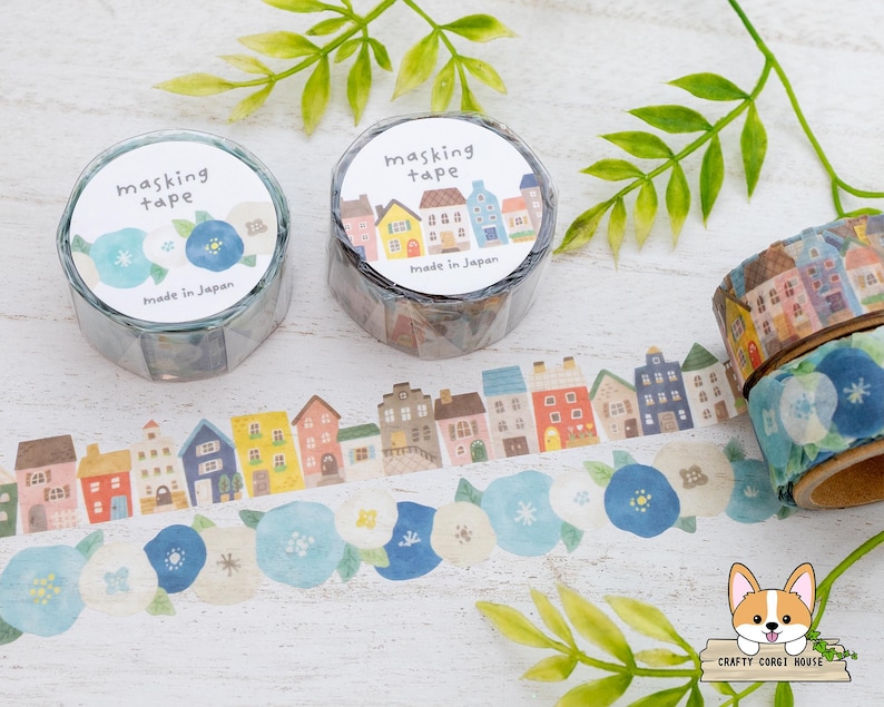May include: Masking tape rolls with floral and house designs. One roll features blue flowers, another has colorful houses. The tape is unrolled, displaying the patterns. Green leaves and the text "masking tape" are also visible.