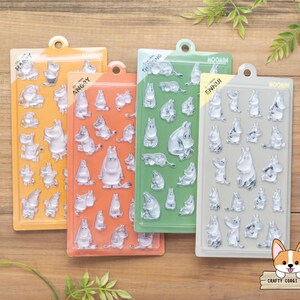 May include: Four rectangular sticker sheets featuring clear Moomin character designs. The sheets are colored orange, coral, green, and gray, each labeled with emotions like HAPPY, ANGRY, and ENNUI. Each sheet contains multiple small, transparent Moomin stickers.