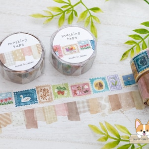 May include: Two rolls of masking tape with "masking tape" printed on the label. One roll has a patterned design, and the other has a stamp design. A strip of tape with various stamp designs is unrolled. The text "Made in Japan" is visible.