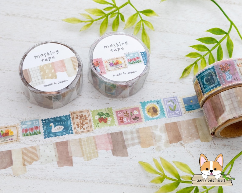 May include: Two rolls of masking tape with "masking tape" printed on the label. One roll has a patterned design, and the other has a stamp design. A strip of tape with various stamp designs is unrolled. The text "Made in Japan" is visible.