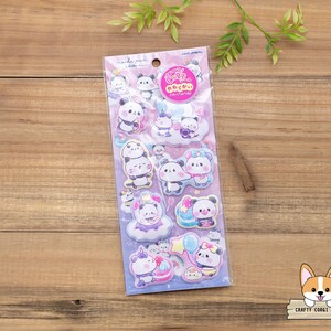 May include: A sheet of stickers featuring cartoon pandas in various poses, some with clouds and balloons. The stickers are in a clear plastic package with Japanese text and a pink logo. The background is a wooden surface.