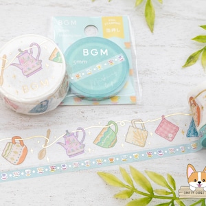 Pode incluir: Um rolo de fita washi com um design de fio dourado que apresenta um bule de chá e xícaras de chá. A fita está sobre uma superfície de madeira branca com dois rolos de fita washi no fundo. A fita está etiquetada "BGM 5mm Morning Tea Time".