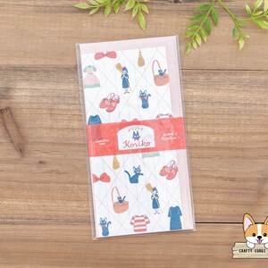 May include: A pink and white envelope with a repeating pattern of a black cat, a red shoe, a broom, and a dress. The envelope has the text "Finy Koriiko Letter & Envelope Set" printed on the front.