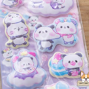 May include: A sheet of colorful stickers featuring cartoon panda bears in various poses. The stickers have a pastel color scheme with pink, blue, and purple accents. Some pandas are wearing crowns or holding treats. The stickers are on a clear backing.