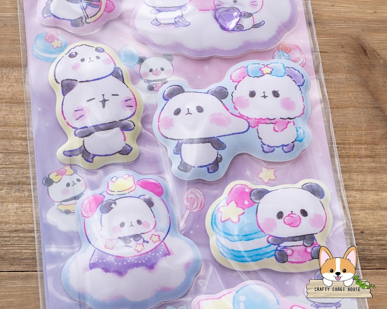 May include: A sheet of colorful stickers featuring cartoon panda bears in various poses. The stickers have a pastel color scheme with pink, blue, and purple accents. Some pandas are wearing crowns or holding treats. The stickers are on a clear backing.