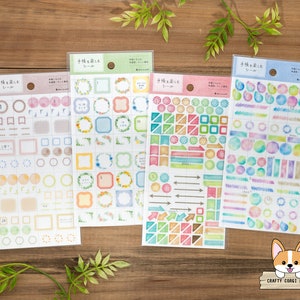 May include: Four sheets of decorative stickers with watercolor designs. The stickers include frames, circles, arrows, and geometric shapes in pastel colors. The text on the packaging is in Japanese. The stickers are on a wooden surface.