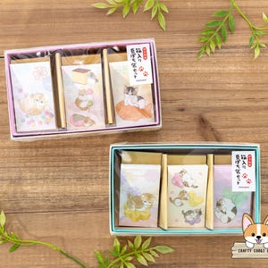 以下が含まれることがあります： 猫をテーマにした文房具セットが入った、2つの装飾的なギフトボックス。各ボックスには、猫のイラストと木製の鉛筆が入った3つのメモ帳が入っています。ボックスはピンクとブルーで、文房具はパステルカラーと日本語のテキストが特徴です。