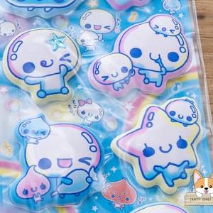 May include: A sheet of colorful stickers featuring cartoon characters with round heads, blue eyes, and smiling faces. The stickers have a pastel color scheme with blue, pink, and yellow accents. Some stickers are shaped like stars and bubbles.