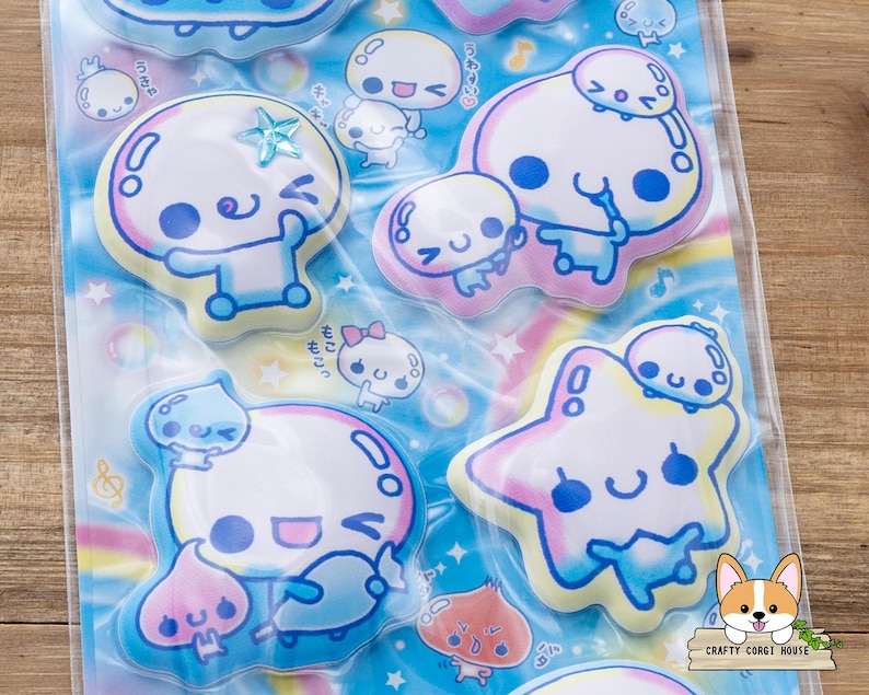 May include: A sheet of colorful stickers featuring cartoon characters with round heads, blue eyes, and smiling faces. The stickers have a pastel color scheme with blue, pink, and yellow accents. Some stickers are shaped like stars and bubbles.