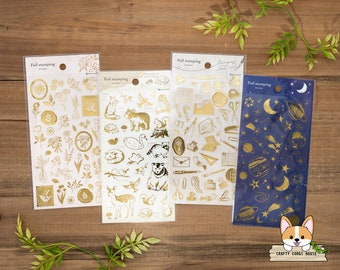 May include: Four sheets of gold foil stickers with various designs. The first sheet features floral and nature-inspired designs. The second sheet features animals and nature-inspired designs. The third sheet features office supplies and stationery designs. The fourth sheet features stars, moons, and planets on a blue background. The text on the sheets reads "Foil stamping sticker".