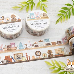 Puede incluir: Dos rollos de cinta adhesiva decorativa con un diseño que presenta osos de peluche, conejitos y varios artículos para el hogar. La cinta está etiquetada como "PAPIER PLATZ Masking tape W20mm x 7m". Una tira de la cinta está desenrollada, mostrando el patrón repetido.