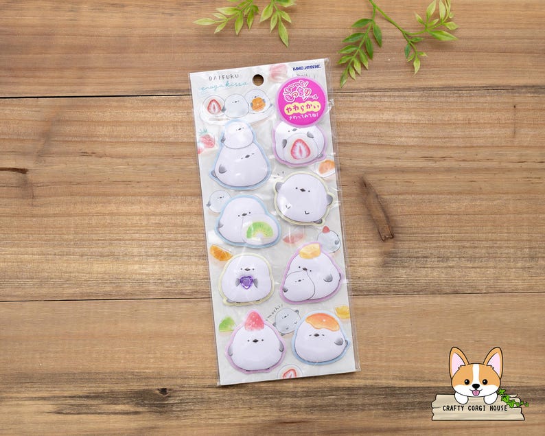 May include: A sheet of stickers featuring cartoon characters resembling mochi desserts. Each sticker has a white base with colorful fruit-themed accents, such as strawberries, oranges, and grapes. The stickers are packaged on a clear backing with Japanese text and a pink circular sticker.