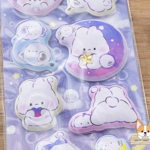 May include: A sheet of stickers featuring cute cartoon characters in various poses. The stickers have a soft color palette of blues, purples, and pinks. Some stickers depict characters on moons or clouds, with star and bubble accents. The stickers are packaged in a clear plastic sheet.