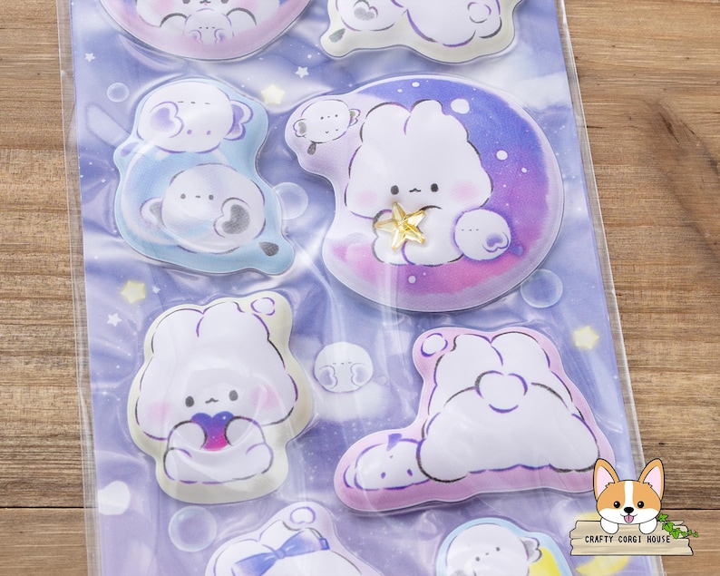 May include: A sheet of stickers featuring cute cartoon characters in various poses. The stickers have a soft color palette of blues, purples, and pinks. Some stickers depict characters on moons or clouds, with star and bubble accents. The stickers are packaged in a clear plastic sheet.