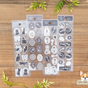 May include: Three sheets of clear sticker paper with black and white illustrations. The first sheet features black cats and bows. The second sheet features keys, locks, and swirls. The third sheet features stars, moons, and other celestial designs.