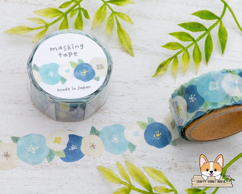 May include: A roll of masking tape with a floral design in shades of blue and white. The tape is in a clear plastic container with the words "masking tape" and "made in Japan" printed on it. The tape is unrolled, showing the floral pattern.