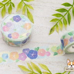 May include: Decorative washi tape featuring a floral design with blue, purple, orange, and yellow flowers. The tape roll has the letters "BGM" printed on it. The tape is unrolled, showing the repeating floral pattern.
