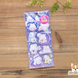 May include: A package of stickers featuring cartoon bunnies in various poses, set against a blue and purple background. The stickers have a cute, whimsical design with soft colors and rounded shapes. The package has the word "fuamii" printed on it.