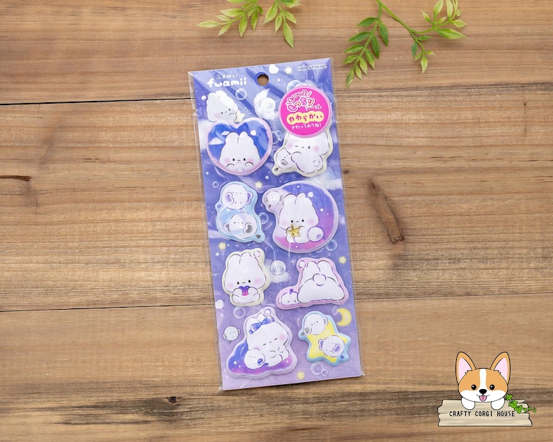 May include: A package of stickers featuring cartoon bunnies in various poses, set against a blue and purple background. The stickers have a cute, whimsical design with soft colors and rounded shapes. The package has the word "fuamii" printed on it.