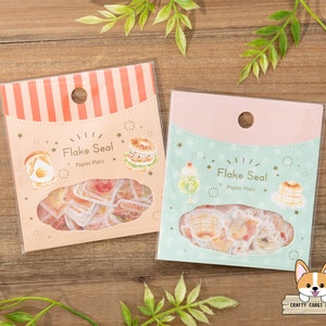 May include: Two packs of colourful paper stickers with a light pink and light blue background. The stickers feature illustrations of sandwiches, cakes, and other food items. The text on the stickers reads "Flake Seal Papier Platz".