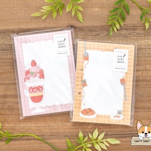 May include: Two pink and orange memo pads with a checkered pattern. The memo pad on the left features a cartoon illustration of a strawberry sundae. The memo pad on the right features a cartoon illustration of a plate of spaghetti. Both memo pads have the text "OTE GARU MEMO" printed on them.