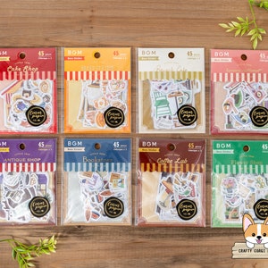 May include: A set of 10 sheets of decorative paper stickers featuring various illustrations of shops and businesses. The stickers are packaged in clear plastic bags with colourful borders and text that reads "BGM" and "Little Shop". The sticker themes include "Cake Shop", "Bread Station", "French Cafe", "Japanese Sweet", "Antique Shop", "Bookstore", "Coffee Lab", and "Flower Shop".