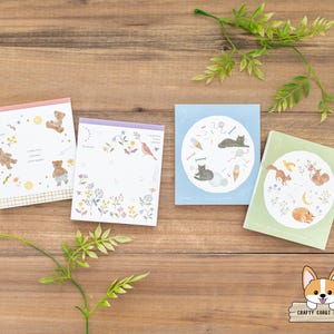 May include: Four colourful notepads with different illustrations. The notepads feature a bear, a cat, a squirrel and a bird. The notepads are on a wooden surface.