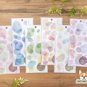 Könnte beinhalten: Sechs Blätter farbiges Aquarell-Stickerpapier mit verschiedenen Formen und Größen von Kreisen und Ovalen. Die Sticker befinden sich auf einem Holzgrund.
