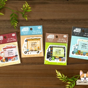 May include: Four colorful, illustrated, stand-up memo pads with a food truck theme. Each pad features a different colored truck with a sign that says "Kotori Machi" and "Welcome!" in Japanese. The pads are 3 inches wide and have a stand-up feature.