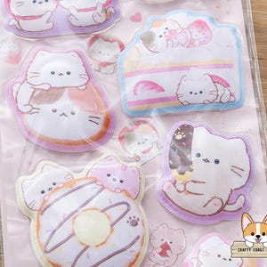 May include: A sheet of cat-themed stickers featuring various designs. The stickers include cats with desserts like cake, donuts, and ice cream. The stickers are in pastel colors with a pink background. The stickers are in a clear plastic package.