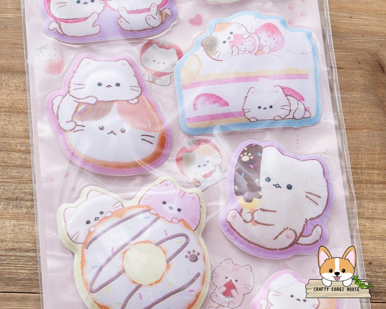 May include: A sheet of cat-themed stickers featuring various designs. The stickers include cats with desserts like cake, donuts, and ice cream. The stickers are in pastel colors with a pink background. The stickers are in a clear plastic package.