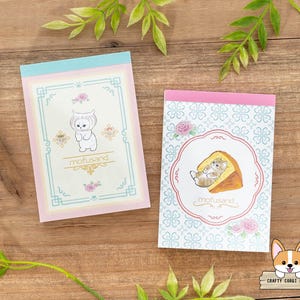 May include: Two pink and blue memo pads with a white border. The top memo pad features a cartoon cat wearing a white hat and the text "mofusand". The bottom memo pad features a cartoon cat laying on a piece of cheese and the text "mofusand".