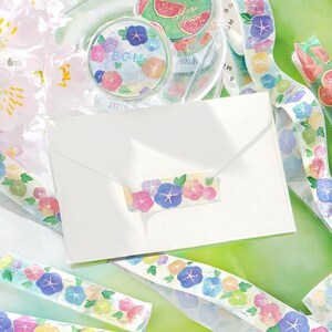 May include: A white envelope with a floral patterned washi tape closure. The tape features blue, pink, and yellow flowers with green leaves. A roll of matching washi tape and watermelon-themed tape are also visible.