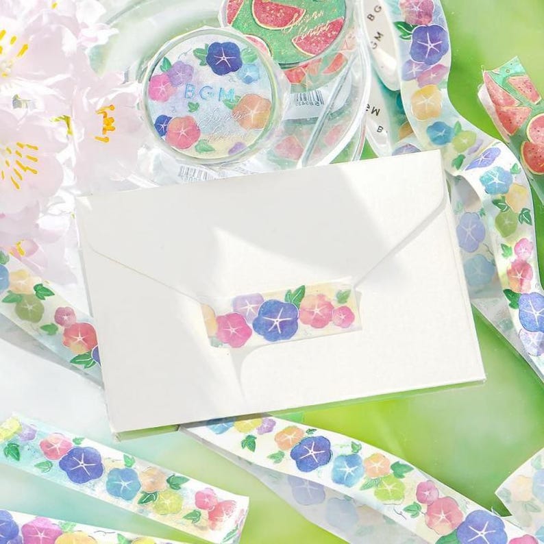 May include: A white envelope with a floral patterned washi tape closure. The tape features blue, pink, and yellow flowers with green leaves. A roll of matching washi tape and watermelon-themed tape are also visible.
