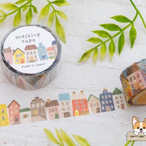 May include: A roll of masking tape with a repeating pattern of colorful houses. The tape is labeled "masking tape" and "Made in Japan". A strip of the tape is unrolled, showing the house design. Green leaves are in the background.