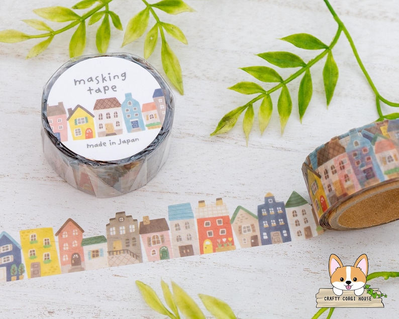 May include: A roll of masking tape with a repeating pattern of colorful houses. The tape is labeled "masking tape" and "Made in Japan". A strip of the tape is unrolled, showing the house design. Green leaves are in the background.