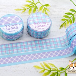 Pode incluir: Dois rolos de fita washi com padr&atilde;o azul e roxo com o texto "きらふわ + SUGAR" e "SUGAR+ BOM Inc." nos rolos.