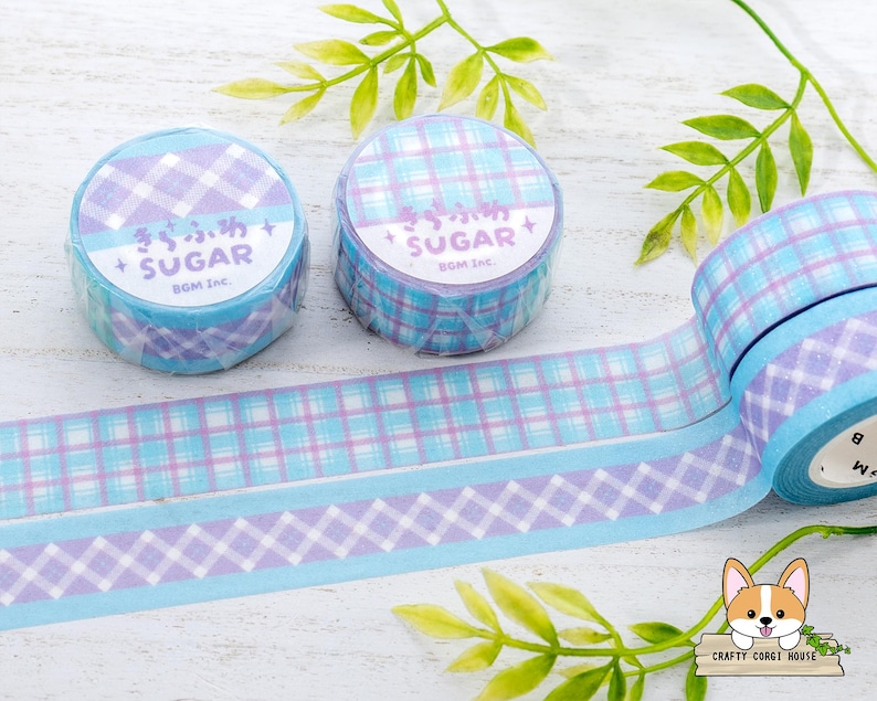Pode incluir: Dois rolos de fita washi com padr&atilde;o azul e roxo com o texto "きらふわ + SUGAR" e "SUGAR+ BOM Inc." nos rolos.