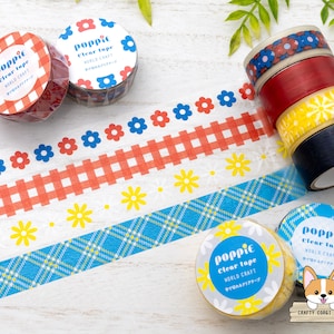 May include: Assortment of colorful washi tapes with floral and geometric patterns. The tapes are rolled up and labeled "poppic Clear Tape" with the text "WORLD CRAFT" and a Japanese phrase. The tapes are displayed on a white wooden surface.