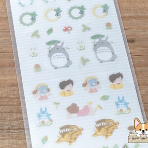 Op de afbeelding: Een vel met transparante stickers met personages en objecten uit de animatiefilm 'Mijn buurman Totoro'. De stickers bevatten Totoro, een kattenbus, een bosgeest en andere grillige ontwerpen. De stickers zijn perfect om een vleugje grilligheid toe te voegen aan dagboeken, planners en andere knutselprojecten.