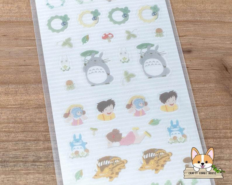 Op de afbeelding: Een vel met transparante stickers met personages en objecten uit de animatiefilm 'Mijn buurman Totoro'. De stickers bevatten Totoro, een kattenbus, een bosgeest en andere grillige ontwerpen. De stickers zijn perfect om een vleugje grilligheid toe te voegen aan dagboeken, planners en andere knutselprojecten.