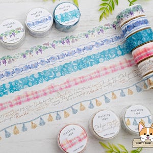 May include: A collection of decorative masking tapes with various designs, including floral patterns, plaid, and tassels. The tapes are in shades of blue, pink, and white, with text labels. Several rolls are stacked, and others are unrolled.