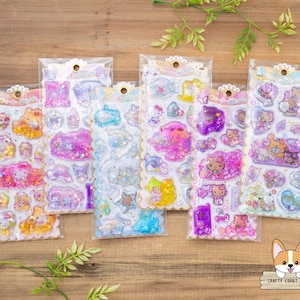 Puede incluir: Una colección de pegatinas brillantes en envases transparentes. Las pegatinas presentan varios personajes de dibujos animados en tonos rosa, morado, azul y amarillo. Las pegatinas tienen un borde festoneado decorativo y están dispuestas sobre una superficie de madera.